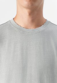 Tæt på en persons hals og øverste bryst, iført en grå t-shirt med ribbet krave og let slidte kanter.