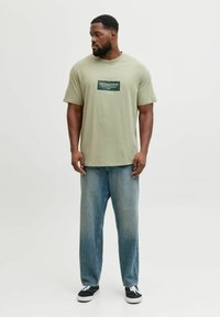 Camiseta de algodón verde claro con logotipo impreso en el pecho, combinada con pantalones vaqueros de mezclilla azul holgados y zapatillas negras. Ajuste casual y ordenado.