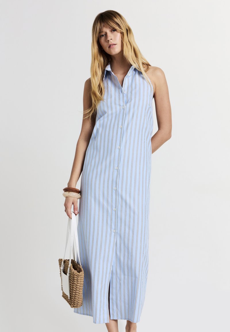 Robe chemise bleu clair avec des rayures verticales marron. Dotée d'un col, d'une fente sur le côté et d'une coupe décontractée. Portée avec un sac en bandoulière tissé.