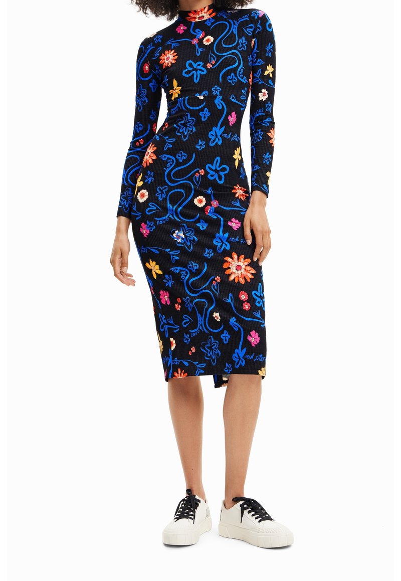 Femme portant une robe noire à manches longues, longueur genou, avec des motifs floraux bleus, roses, orange et jaunes, et des baskets blanches avec des lacets noirs.