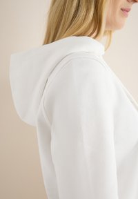 Felpa bianca con una texture morbida, caratterizzata da un cappuccio prominente e cuciture dettagliate lungo le spalle e l'area della zip.