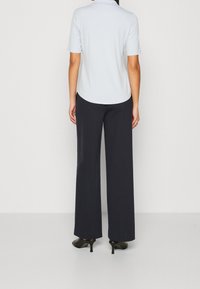 Una donna indossa una camicia a maniche corte blu chiaro, pantaloni neri a gamba larga e scarpe con tacco nere, in piedi di fronte a uno sfondo neutro.