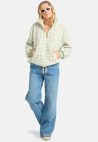 A woman in a light green checked fleece pullover stands confidently with hands in pockets, wearing blue jeans and stylish white sunglasses.