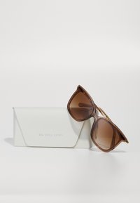 Ochelari de soare maro supradimensionați cu o formă geometrică și lentile gradient, așezati pe un etui texturat alb cu logo-ul "Michael Kors" embosat.