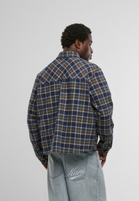 Férfi kék és olivazöld kockás inget és világoskék farmert visel, a hátsó zsebében "Kani" hímzéssel, egy egyszínű háttér előtt áll.