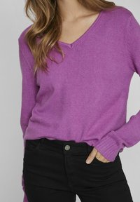 Pull à col en V et manches longues en maille violette vif, associé à un jean noir. Le pull présente une coupe décontractée et des poignets côtelés.