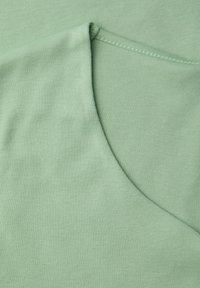 Tela verde claro con una textura suave, que presenta un cuello redondeado y un dobladillo cosido. El color parece uniforme, sin patrones ni acentos.