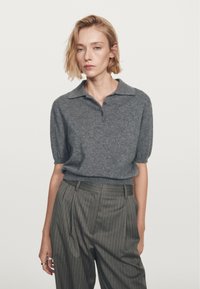 Donna con capelli biondi corti che indossa un polo a maniche corte grigio in maglia e pantaloni a vita alta grigi con righe verticali, in piedi contro uno sfondo bianco.