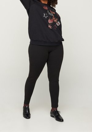 Personne portant des leggings noirs à motifs, un pull festif noir avec des ornements en sequins, des chaussettes bordeaux et des chaussures noires à lacets sur un fond uni.
