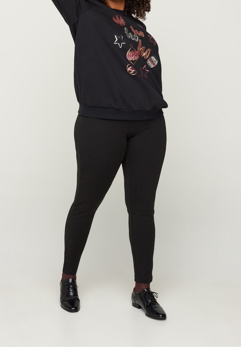 Personne portant des leggings noirs à motifs, un pull festif noir avec des ornements en sequins, des chaussettes bordeaux et des chaussures noires à lacets sur un fond uni.