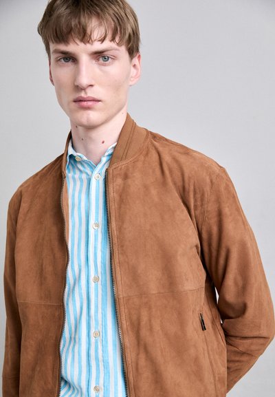 Hnědá semišová bomber bunda se zapínáním na zip, v kombinaci s modro-bílou pruhovanou košilí s knoflíky. Hladká textura a minimální detaily šití.