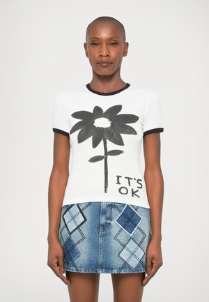 Vrouw in een witte t-shirt met grote zwarte bloem en de tekst "IT'S OK", gecombineerd met een blauwe denim minirok met diamanten patches.