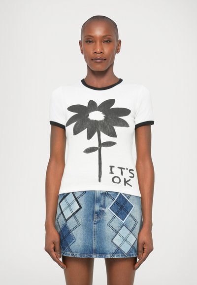 Γυναίκα που φοράει λευκό t-shirt με μεγάλη μαύρη λουλούδα και κείμενο "ΕΙΝΑΙ ΕΝΤΑΞΕΙ", συνδυασμένο με μπλε τζιν μίνι φούστα με διαμαντένια διακοσμητικά.