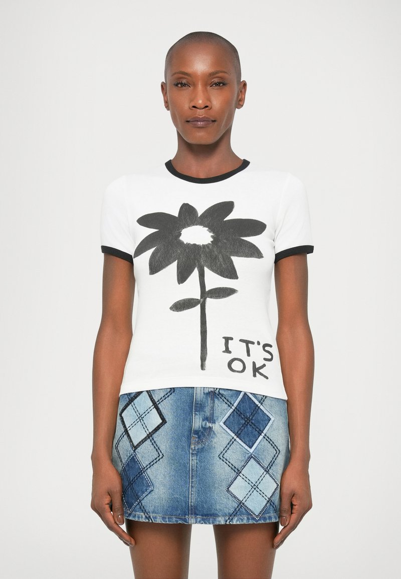 Kvindelig iført en hvid t-shirt med stor sort blomst og teksten "DET ER OK", parret med en blå denim mini-nederdel med diamantlapper.