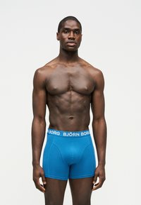 Μυώδης άνδρας με σκουρόχρωμο δέρμα, όρθιος και στραμμένος μπροστά, φορώντας μπλε μποξεράκια Björn Borg, σε απλό φωτεινό φόντο.