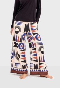 Wide-leg pants in cream fabric, featuring a bold geometric pattern in blue, red, and black, with a striped hem. Lightweight and breathable.