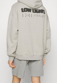 Sweat à capuche gris avec cordon de serrage, arborant "LOW LIGHTS" en peinture noire en spray au dos, associé à un short gris.