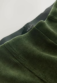 Tissu vert côtelé avec un motif texturé et une taille noire ornée d'une étiquette. Texture lisse avec des lignes verticales et des coutures visibles.