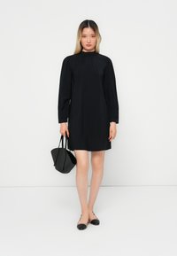 Abito corto nero con maniche lunghe, colletto alto, texture liscia, abbinato a una piccola borsa nera e ballerine nere.