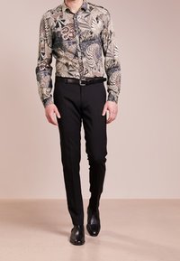 Langarmhemd mit Muster in Beigetönen, Grün und Schwarz, kombiniert mit enganliegenden schwarzen Hosen und schwarzen Lederschuhen.