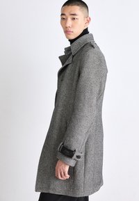 Grå hundtandsmönstrad kappa med en strukturerad krage, sidfickor och knappdetaljer. Tillverkad av texturerat tyg med en skräddarsydd passform.