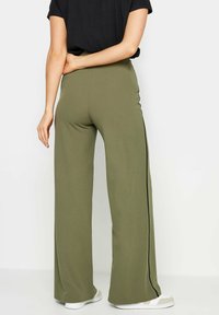Pantaloni a gamba larga verde oliva realizzati in tessuto morbido, con una striscia laterale nera e un design a vita alta. Abbinati a sneakers bianche.