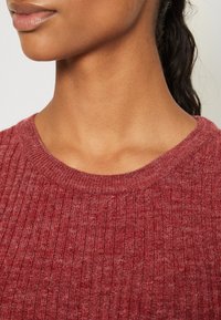 Roter gerippter Pullover mit rundem Ausschnitt, strukturiertem Stoff und leichtem Glanz. Nahaufnahme des Hals- und Schulterbereichs.