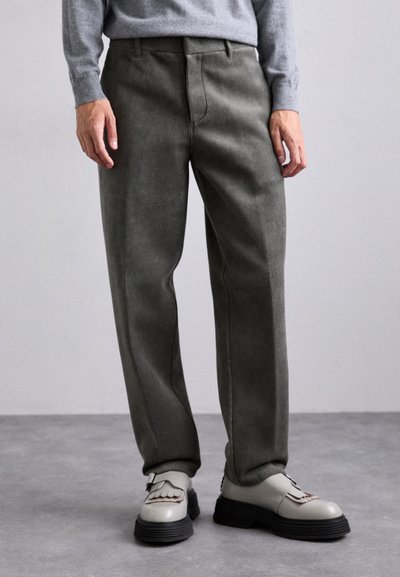 Pantaloni in corduroy verde oliva con taglio dritto, dotati di tasche frontali e superficie testurizzata. Abbinati a scarpe beige con suole scure.