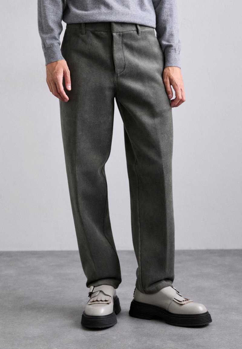 Pantaloni in corduroy verde oliva con taglio dritto, dotati di tasche frontali e superficie testurizzata. Abbinati a scarpe beige con suole scure.