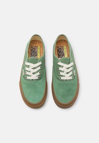Baskets basses en daim vert avec lacets blancs, semelles en caoutchouc gomme, et une semelle intérieure en liège arborant le logo. Texture lisse et design classique.