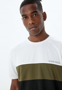 Camiseta de manga corta en blanco, verde oliva y negro. Texto "NO MÁS EXCUSAS" en gris en la parte frontal. Tejido de algodón suave, diseño de cuello redondo.