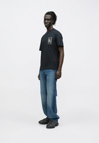 Άνδρας με ντραντλόκς, φορώντας μαύρο μπλουζάκι με σχέδιο, φαρδιά μπλε τζιν και μαύρα αθλητικά, στέκεται μπροστά σε λευκό απλό φόντο.