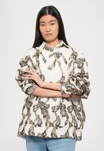 Oversized beige sweater with black and gold floral embroidery, wide sleeves, and a round neckline. Worn over a white collared shirt.