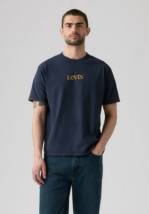 Mężczyzna o krótkich włosach i brodzie nosi ciemnoniebieską koszulkę Levi's i niebieskie dżinsy, stoi na tle jednolitego jasnoszarego tła.