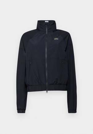 Viegls melns vējjaka ar augstu apkakli, priekšējo rāvējslēdzēju un elastīgām aprocēm; uz krūtīm ir neliels zaļš logs.