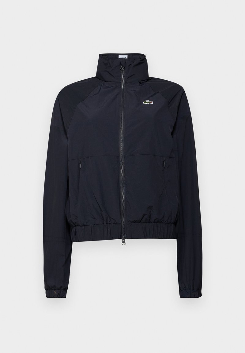 Veste coupe-vent noire légère avec un col haut, fermeture éclair frontale et poignets élastiques ; comporte un petit logo vert sur la poitrine.