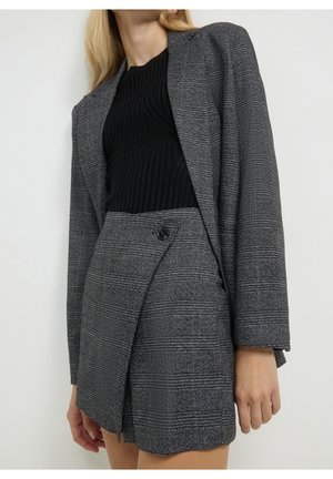 Femme portant un blazer à carreaux gris et une mini-jupe taille haute assortie avec un pull côtelé noir sur un fond uni.