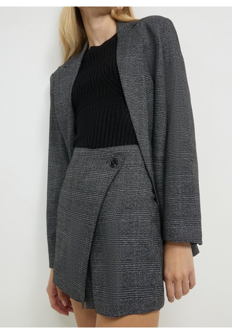 Femme portant un blazer à carreaux gris et une mini-jupe taille haute assortie avec un pull côtelé noir sur un fond uni.