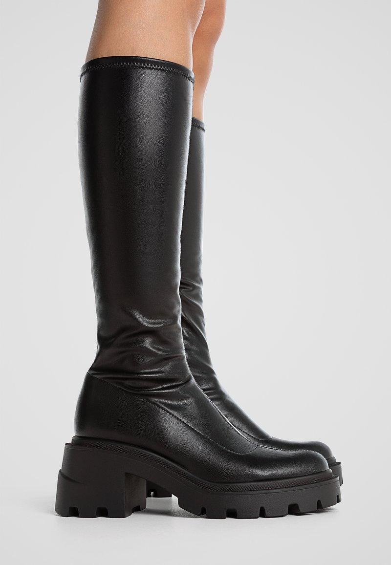 Bottes en cuir noir hauteur genou avec un design élégant, une forme courbée, une finition texturée et une semelle à crampons épaisse pour plus d'adhérence et de stabilité.