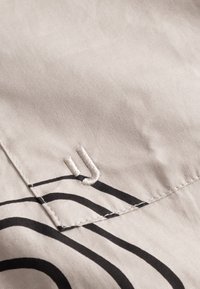 Világos bézs színű pamut szövet fekete spirálmintákkal. Jellemzője egy varrott "U" betű és látható varrási részletek.