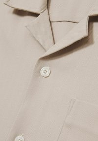 Camisa de botones de espiga beige con cuello notched, que cuenta con un bolsillo en el pecho y un detalle de botón blanco redondo.