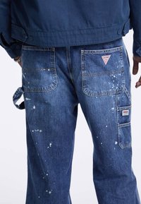 Pantaloni cargo in denim blu con macchie di vernice, numerose tasche e una toppa con logo sul retro. Design a vestibilità casual e strutturata.