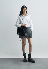 Chemise blanche à boutons, jupe en cuir noire avec fermeture éclair, sac à main noir à franges, et bottes noires à lacets avec œillets dorés. Fond gris.