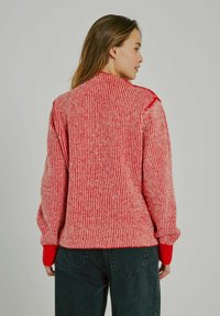 Maglione a righe rosse e crema con texture a coste, dotato di maniche lunghe e polsini rossi a tinta unita, abbinato a jeans scuri.