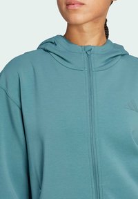 Svijetlo plavi zip-up dukserica s kapuljačom, koja ima glatku teksturu i suptilno brendiranje na prsima. Zatvaranje sprijeda na zip, ležeran kroj.