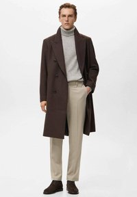 Manteau en laine marron à double boutonnage, col roulé côtelé gris, pantalon beige cintré et chaussures marron foncé. Lignes épurées et design structuré.