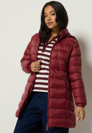 Femme portant une longue veste matelassée bordeaux par-dessus un polo rayé et un pantalon bleu, debout devant un fond clair uni.