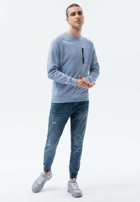 Ljusblå sweatshirt med en dragkedja ficka, matchad med slitna blå jeans och grå sneakers med vita snörningar. Avslappnad passform och design.