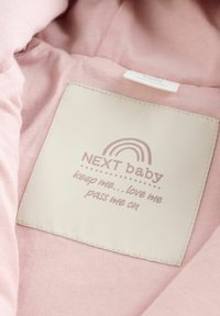 Lys rosa stof med en rektangulær creme etiket, der viser "NEXT baby" og tekst nedenunder, præget regnbue design og syede kanter.