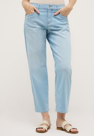 Hellblaue Jeans mit geradem Bein, getragen mit beigen flachen Sandalen, Hände in den Taschen ruhend, stehend vor schlichtem hellem Hintergrund.